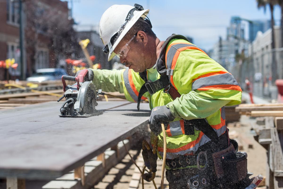 Construction worker in safety gear cutting plywood at an urban transit construction site.