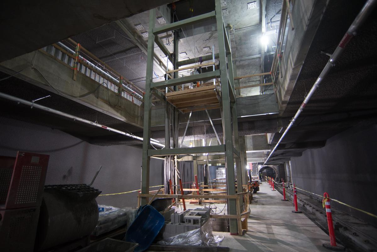 Interior construction of transit station with green steel framework, elevator installation equipment, and LED lighting along concrete platform.