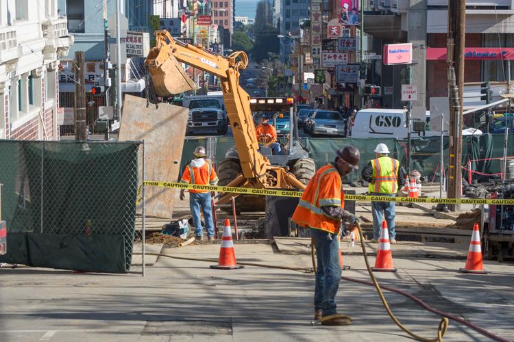 Construction crew operating yellow excavator on urban street with traffic cones and caution tape, surrounded by buildings and city infrastructure.