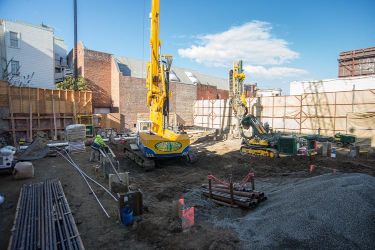 Construction site with yellow drilling equipment and excavation machinery on urban lot surrounded by brick buildings.