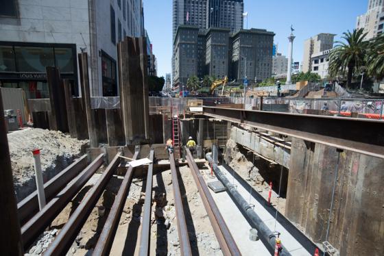 Construction site showing steel beams and wooden shoring at urban street intersection with city skyline.