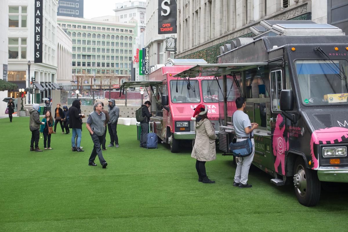 Food trucks parked on green lawn in downtown plaza with pedestrians and buildings in background.