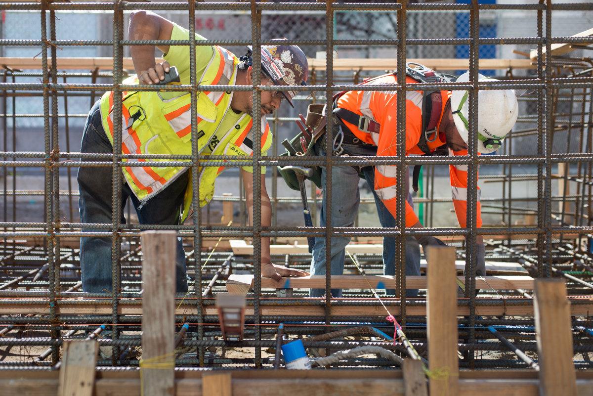 Two construction workers in safety vests installing rebar at a transit station construction site.