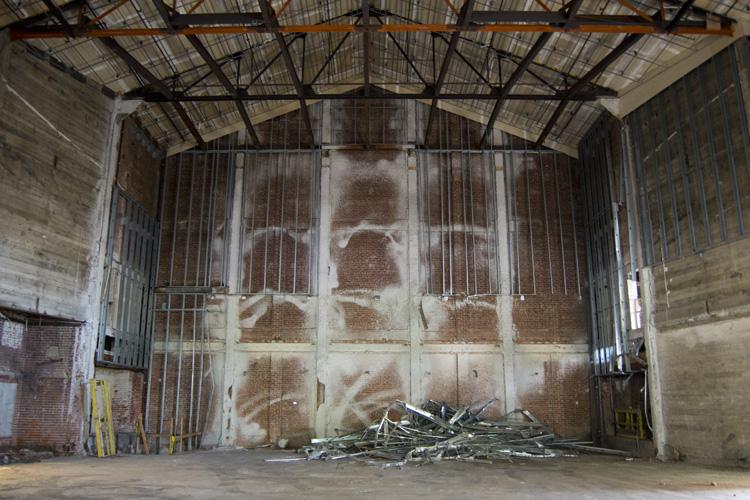 Interior of abandoned warehouse with exposed brick walls, metal framework ceiling, and debris pile on floor.