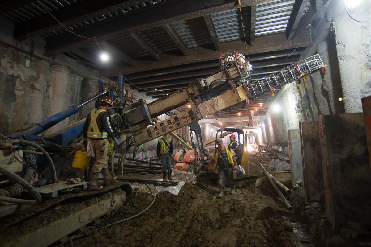 Underground station excavation with temporary bracing, drilling equipment, and workers in hard hats at construction site.