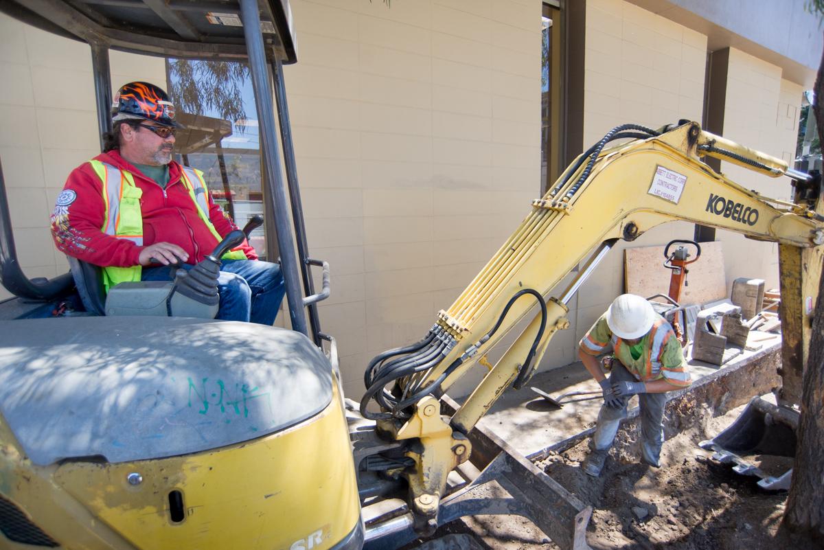 Construction workers operate yellow excavator during Central Subway sidewalk restoration and utility work.