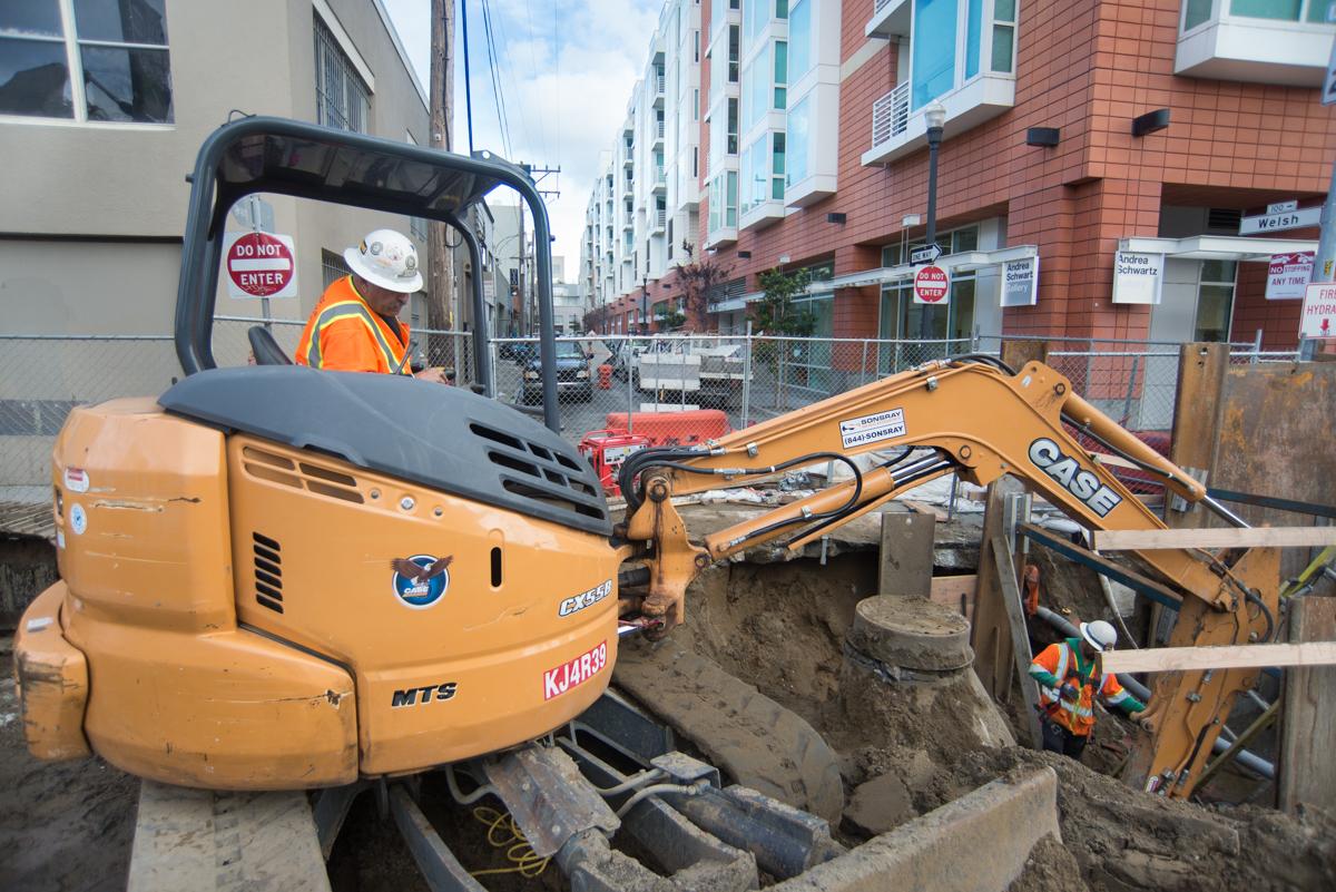 Construction excavator removing soil at 4th Street work site with operator in safety gear and residential buildings in background.