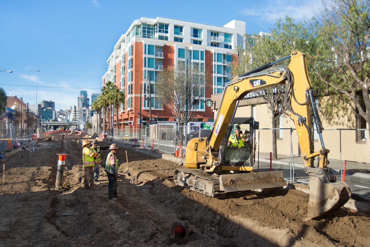 Construction crew operates yellow excavator at 4th and Brannan station site with modern residential building in background.