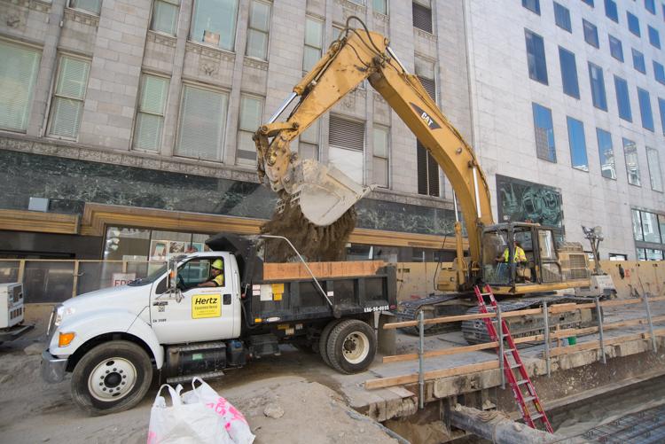 Excavator with slotted bucket loading debris into Hertz dump truck at Union Square Garage construction site.