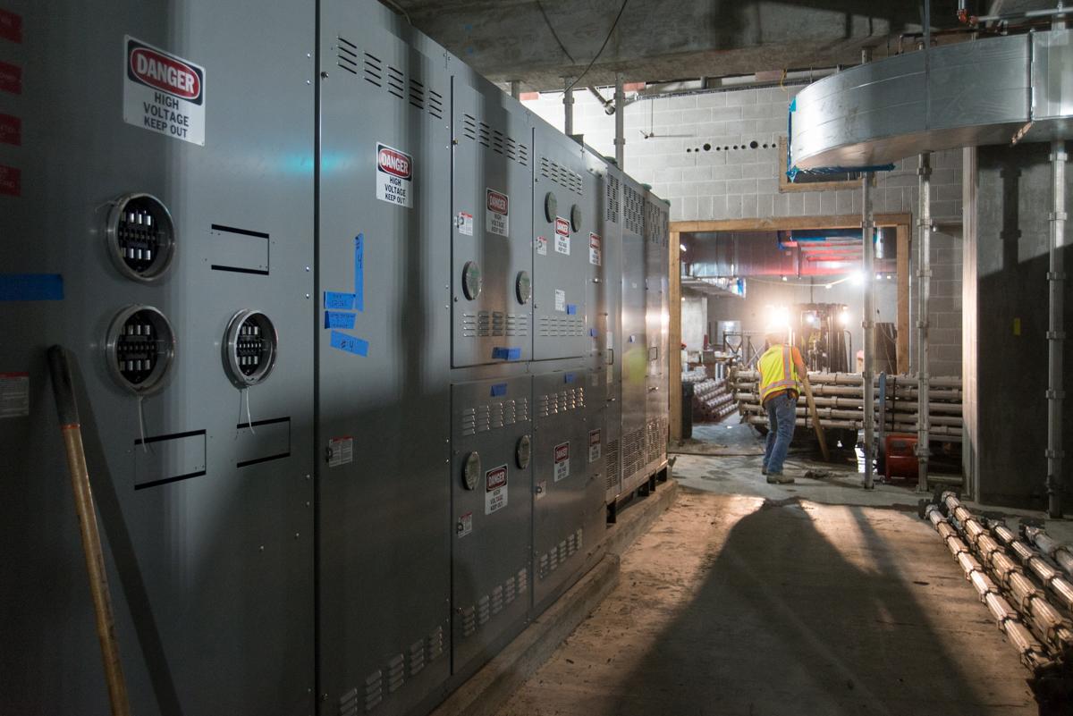 Electrical equipment and control panels line a transit station corridor during construction, with a worker in safety gear visible in the background.