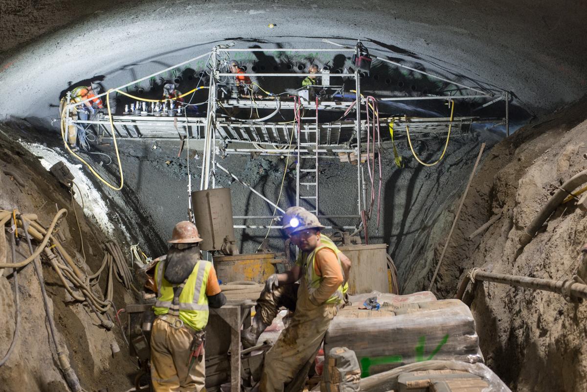 Workers in safety gear excavate underground tunnel cavern with drilling equipment and scaffolding visible above.