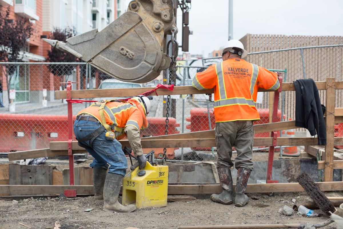 Two construction workers in orange safety vests inspect equipment at a trench during Central Subway utility installation work.