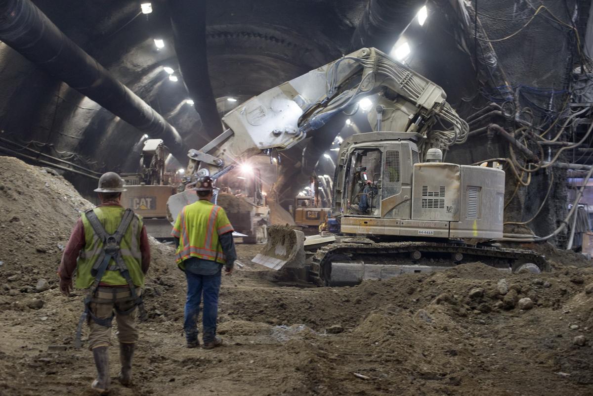 Workers in safety vests monitor excavation equipment breaking apart concrete in an underground transit tunnel.