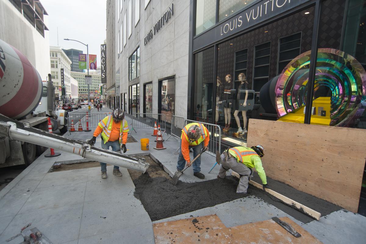 Construction workers in safety vests pouring concrete on an urban street next to a Louis Vuitton storefront.