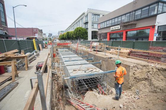 Construction worker in safety gear at Central Subway 4th Street Station excavation site with reinforced concrete foundation work.