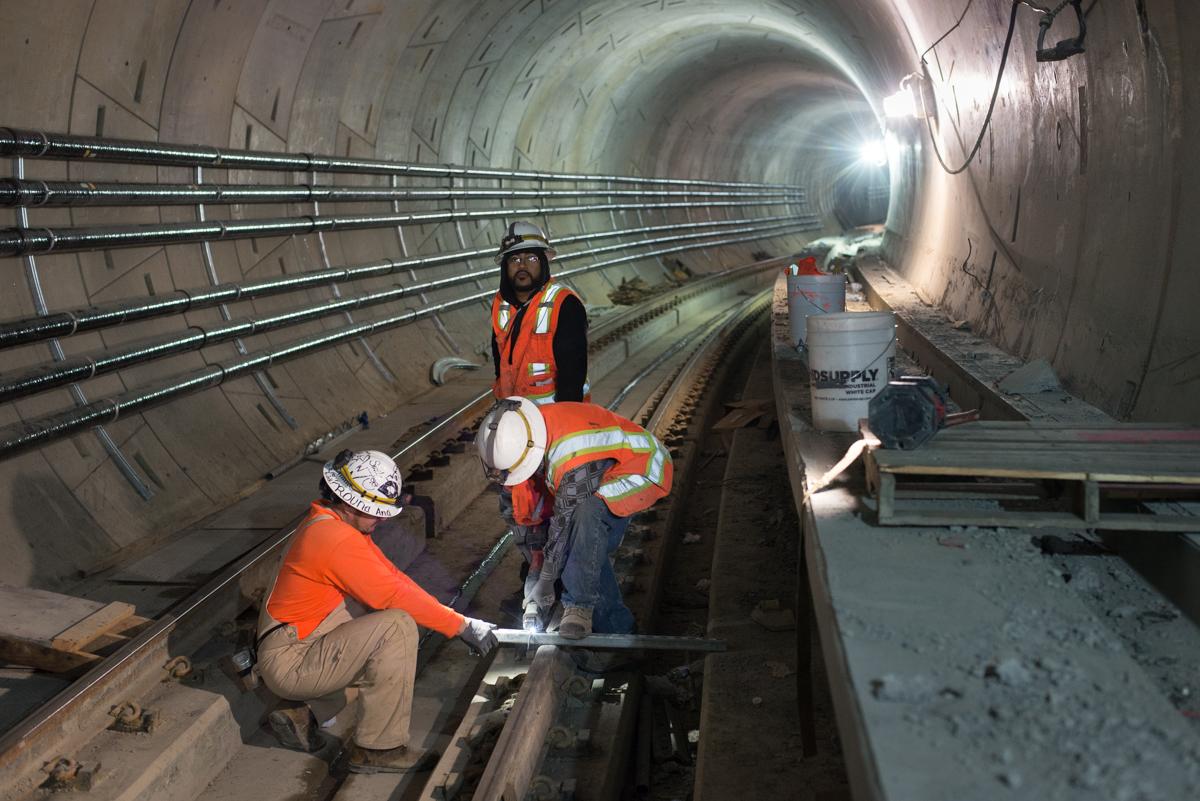 Workers in hard hats and safety vests install mounting brackets inside a subway tunnel under construction.