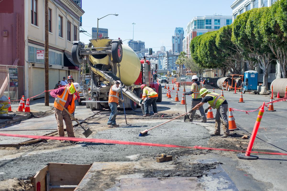 Construction workers operate a concrete mixer truck during sewer utility work on an urban street.