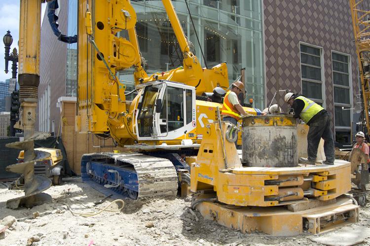Construction crew operates yellow excavator on urban street with concrete barriers and safety equipment visible.