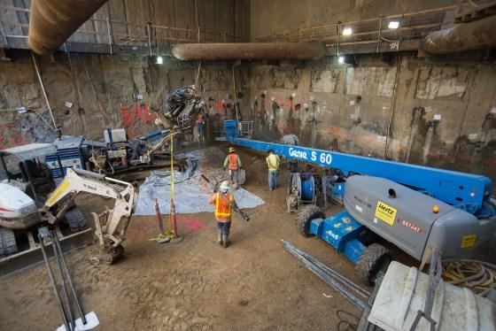 Construction workers in safety gear operate machinery inside underground transit station during waterproofing repairs.