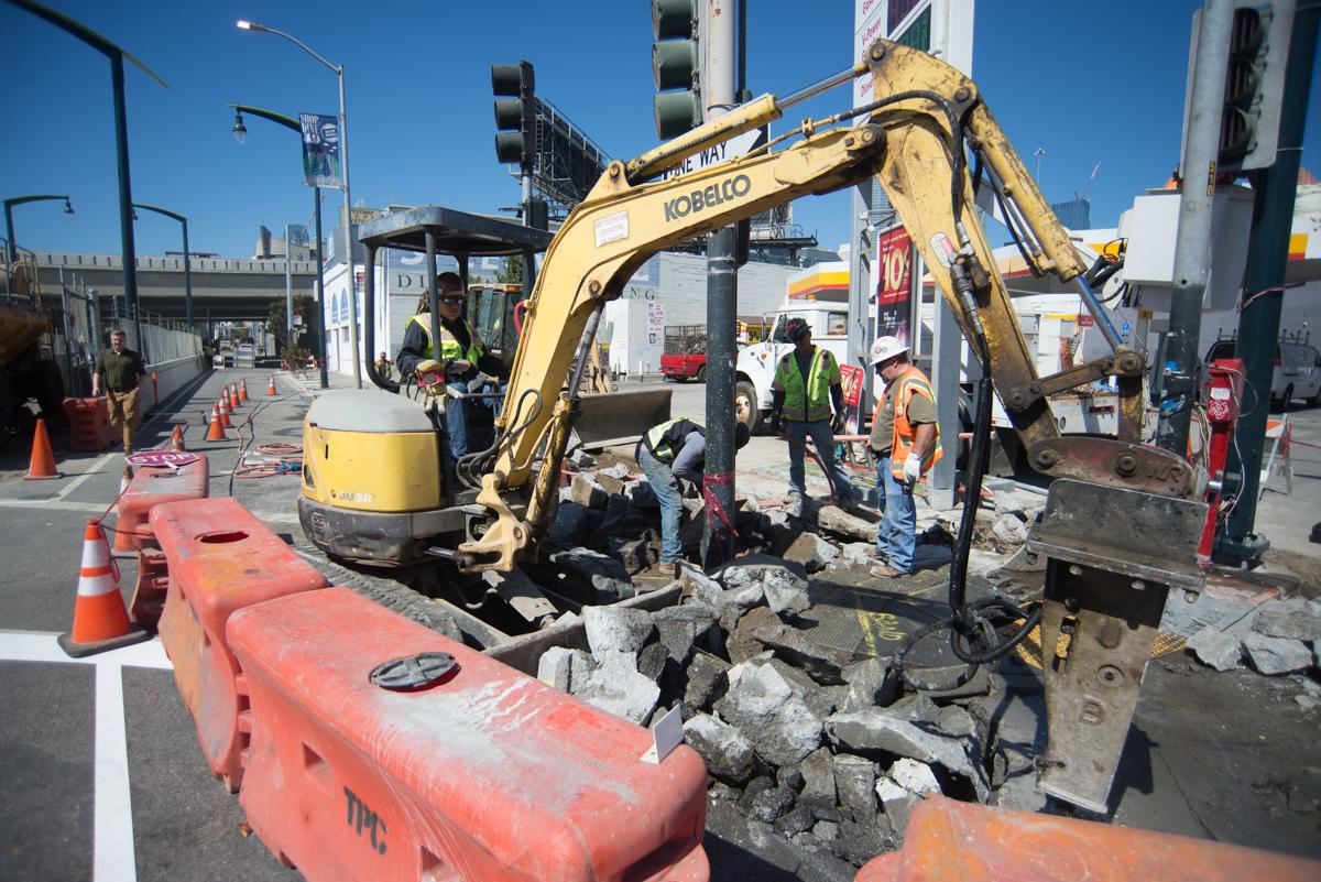 Kobelco excavator removes concrete debris at 4th and Brannan construction site with workers and traffic control barriers present.