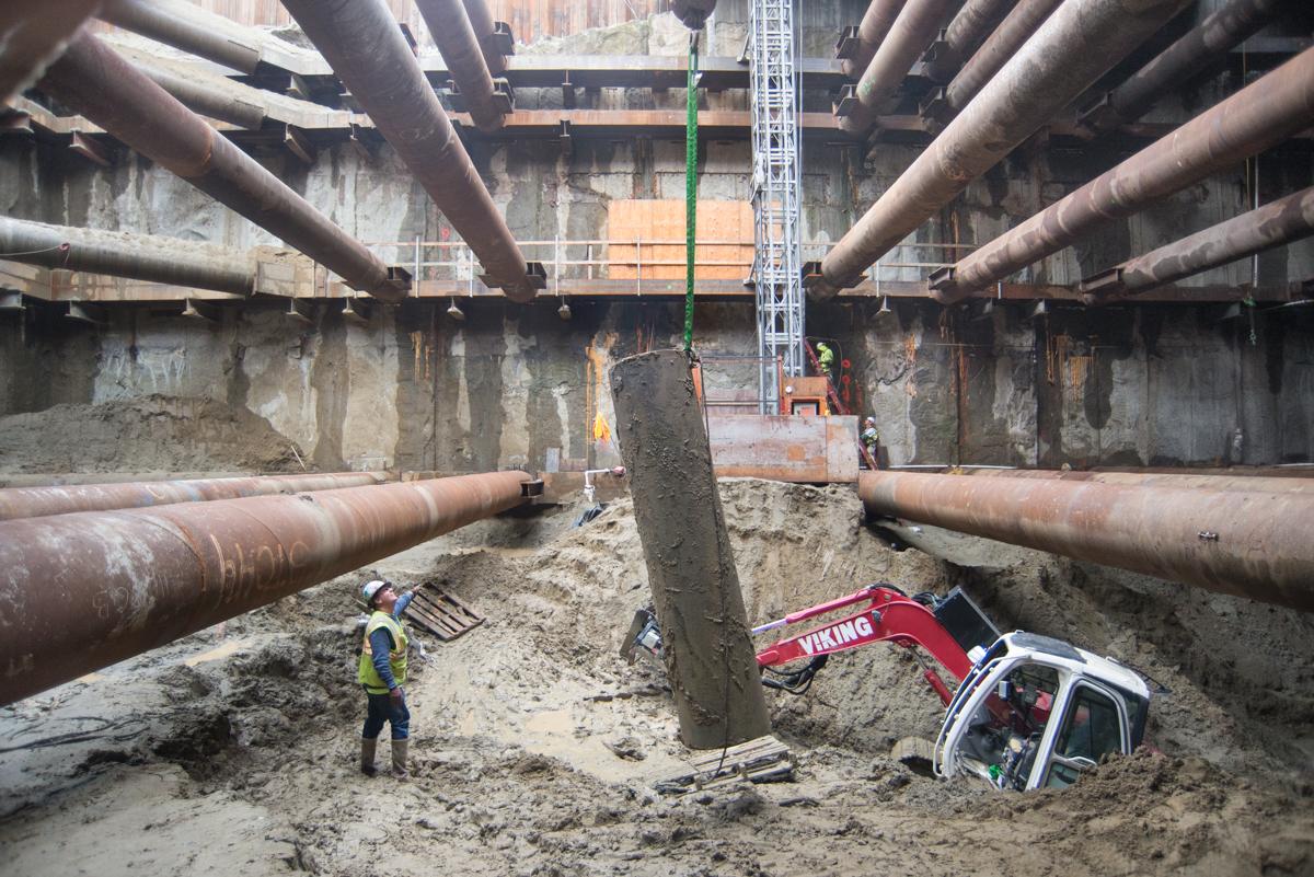 Construction site showing excavation pit with large steel pipes overhead, red excavator, and workers in safety gear beneath concrete infrastructure.