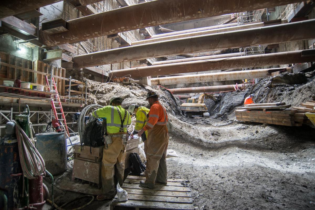 Workers in safety vests examine equipment inside an underground construction site with steel beams and excavated material.