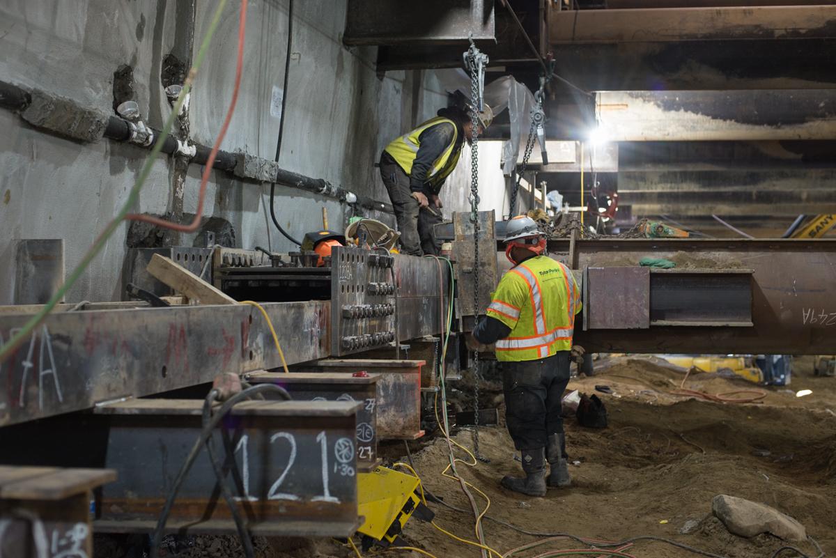 Workers in safety vests install temporary bracing and struts inside an underground station box construction site.
