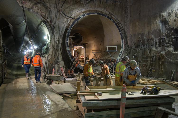 Workers in safety gear construct tunnel sections beneath 4th Street, with large circular tunnel boring equipment visible in underground construction site.