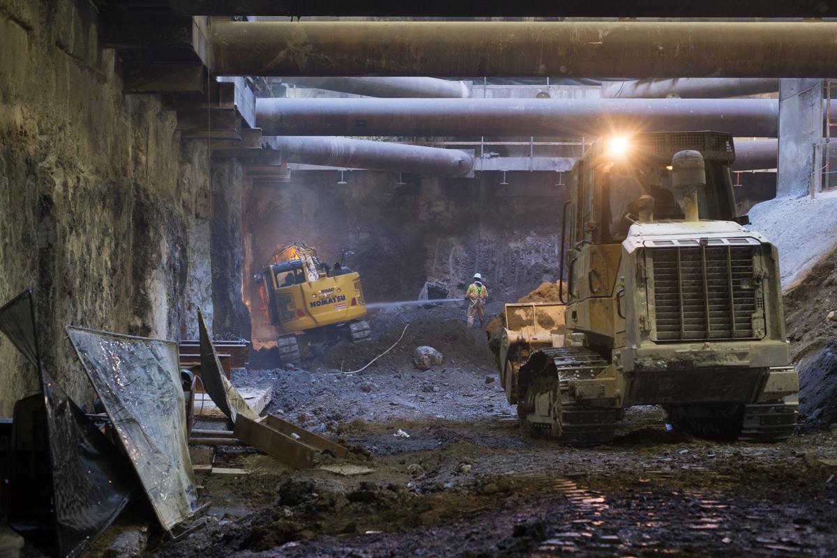 Underground construction site with excavator and dump truck removing concrete debris beneath concrete beams.