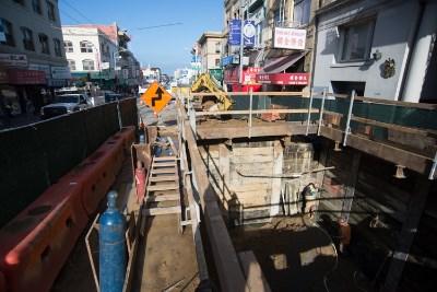 Construction site showing excavation pit with temporary soil supports, heavy equipment, and surrounding urban buildings.