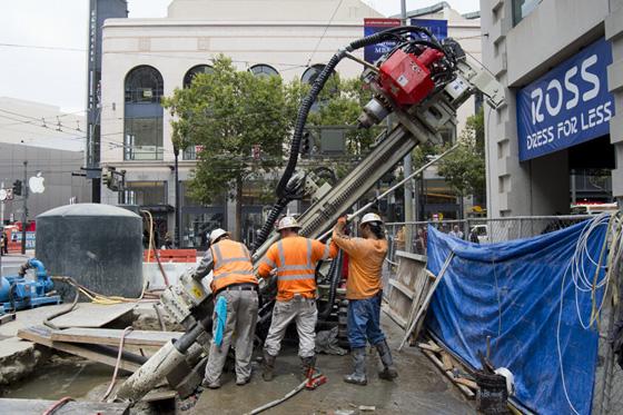 Workers in orange vests operate ground stabilization equipment on a city street surrounded by construction barriers and buildings.