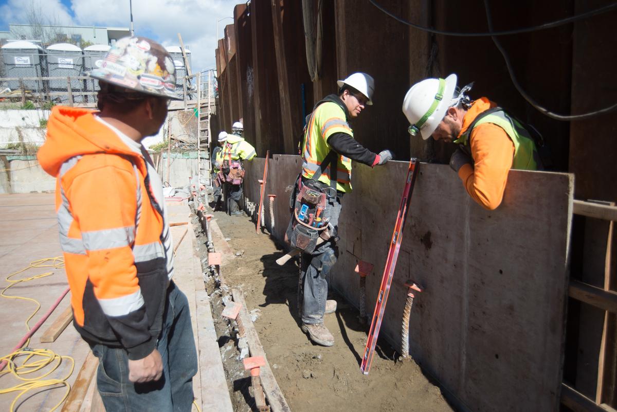 Construction workers in safety gear install machinery and cabling inside a transit station box.