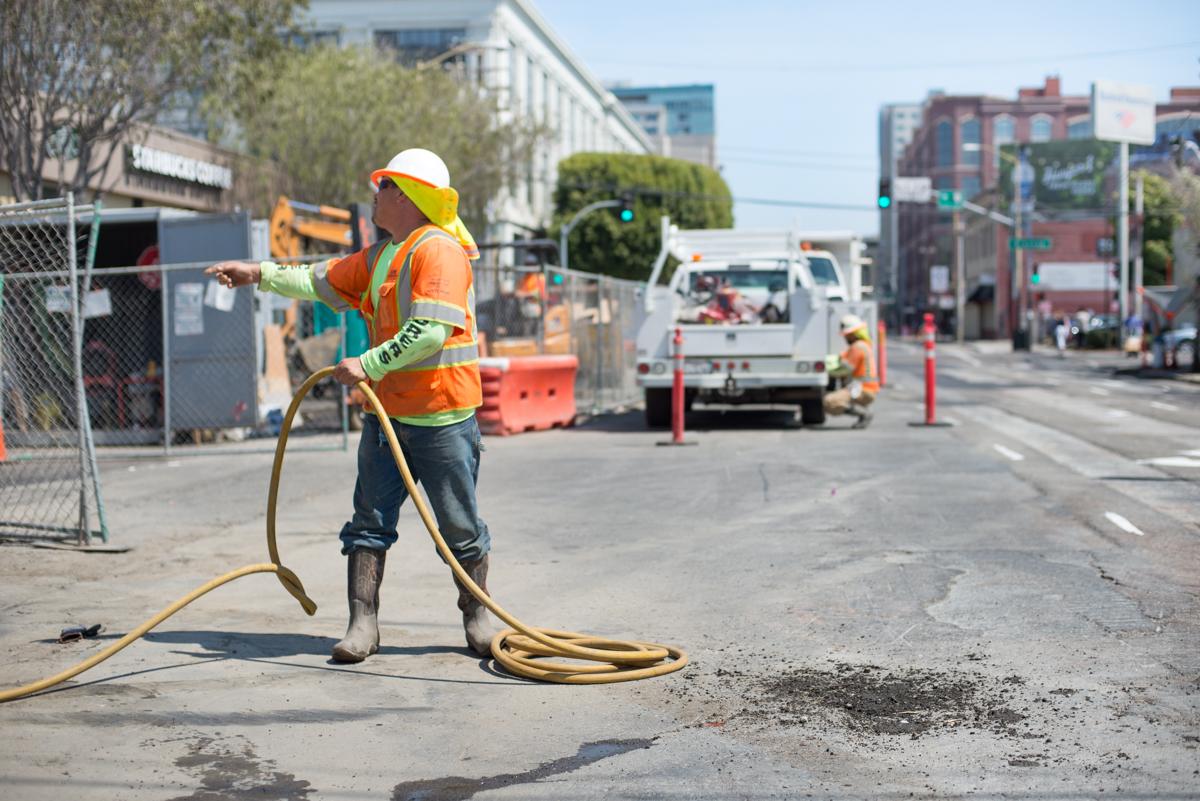 Construction worker in safety gear handling yellow hose at urban work site with equipment and vehicles.
