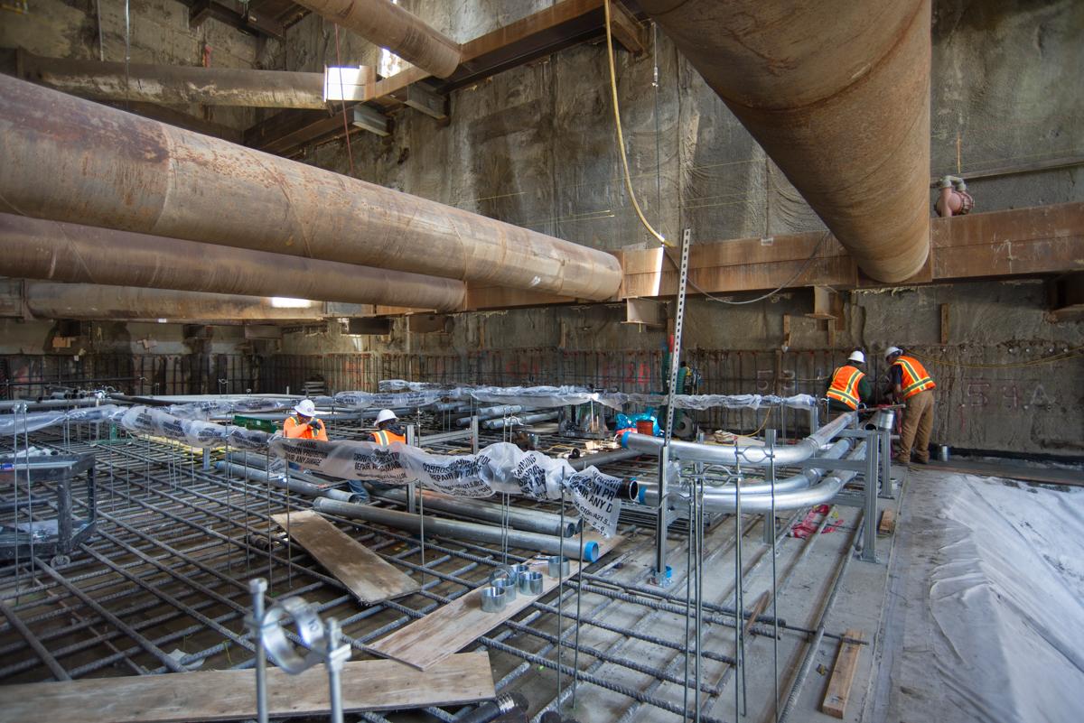 Construction workers install rebar and utilities inside concrete headhouse structure with large pipes overhead.