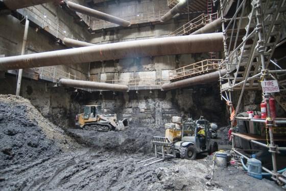 Construction site with excavation equipment, large pipes, and temporary steel supports in underground transit station headhouse.