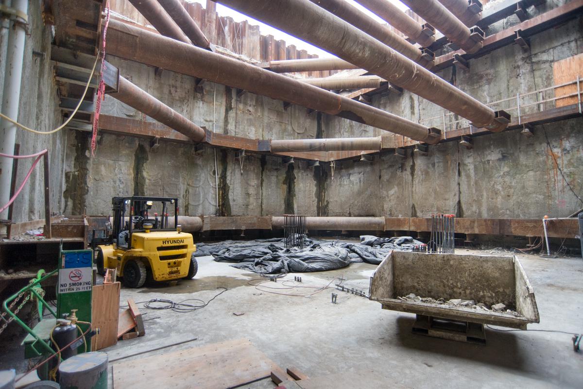 Construction site inside station box with yellow forklift, concrete floor, steel pipes overhead, and temporary bracing removal in progress.