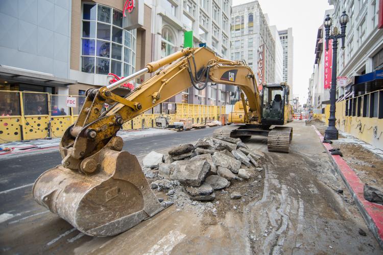 Excavator with bucket attachment removing broken pavement on urban street surrounded by yellow barriers and construction equipment.