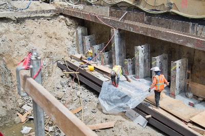 Construction workers installing structural piles at an underground transit project site with protective barriers and equipment.