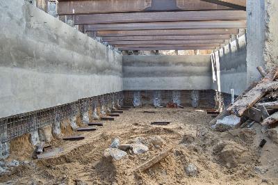 Underground garage construction showing concrete walls, steel beams, and sandy floor during utility relocation work.