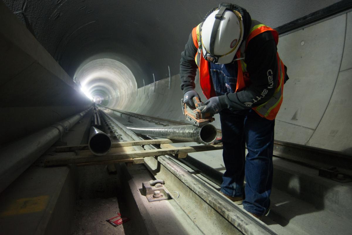 Worker in hard hat and safety vest installing conduit in illuminated underground tunnel.