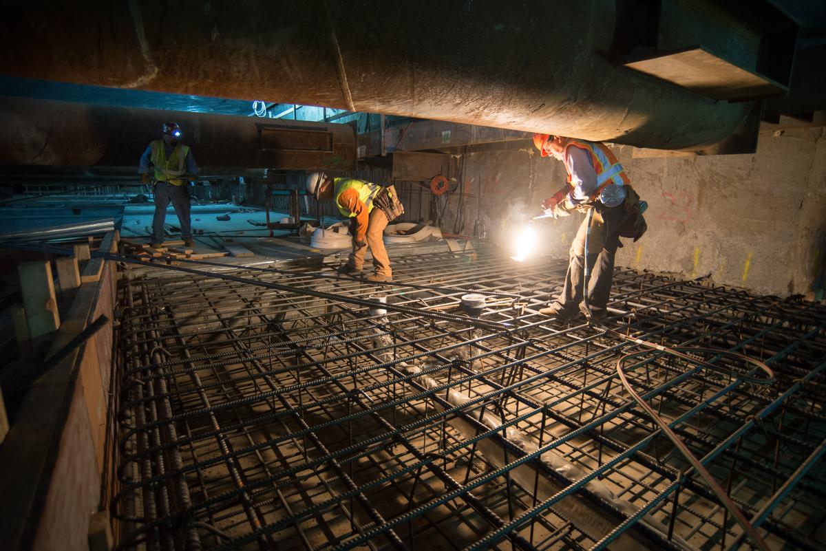 Workers install rebar and construct concrete forms for floor slab during underground transit construction at night.