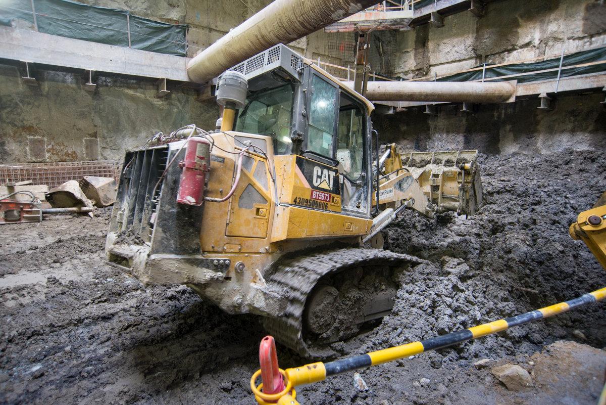 Caterpillar bulldozer moving soil in underground construction site with concrete walls and large pipes overhead.