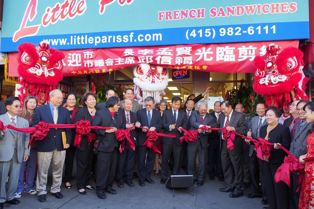 Group of officials and community members at ribbon-cutting ceremony for Little Paris French Sandwiches restaurant in Chinatown, with red decorative lions and bows.