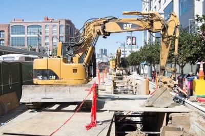 Yellow excavator working on sewer line upgrade at 4th and King Street with open trench and construction barriers.