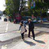 Two people walking on a sidewalk near a street with parked cars and trees.