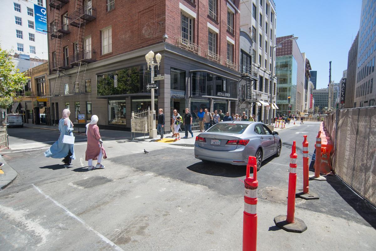 Street intersection with red and white traffic cones blocking lanes, pedestrians walking, and brick buildings lining the urban street.