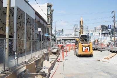 Construction site on 4th Street with yellow excavator, orange safety barriers, and San Francisco skyline in background.