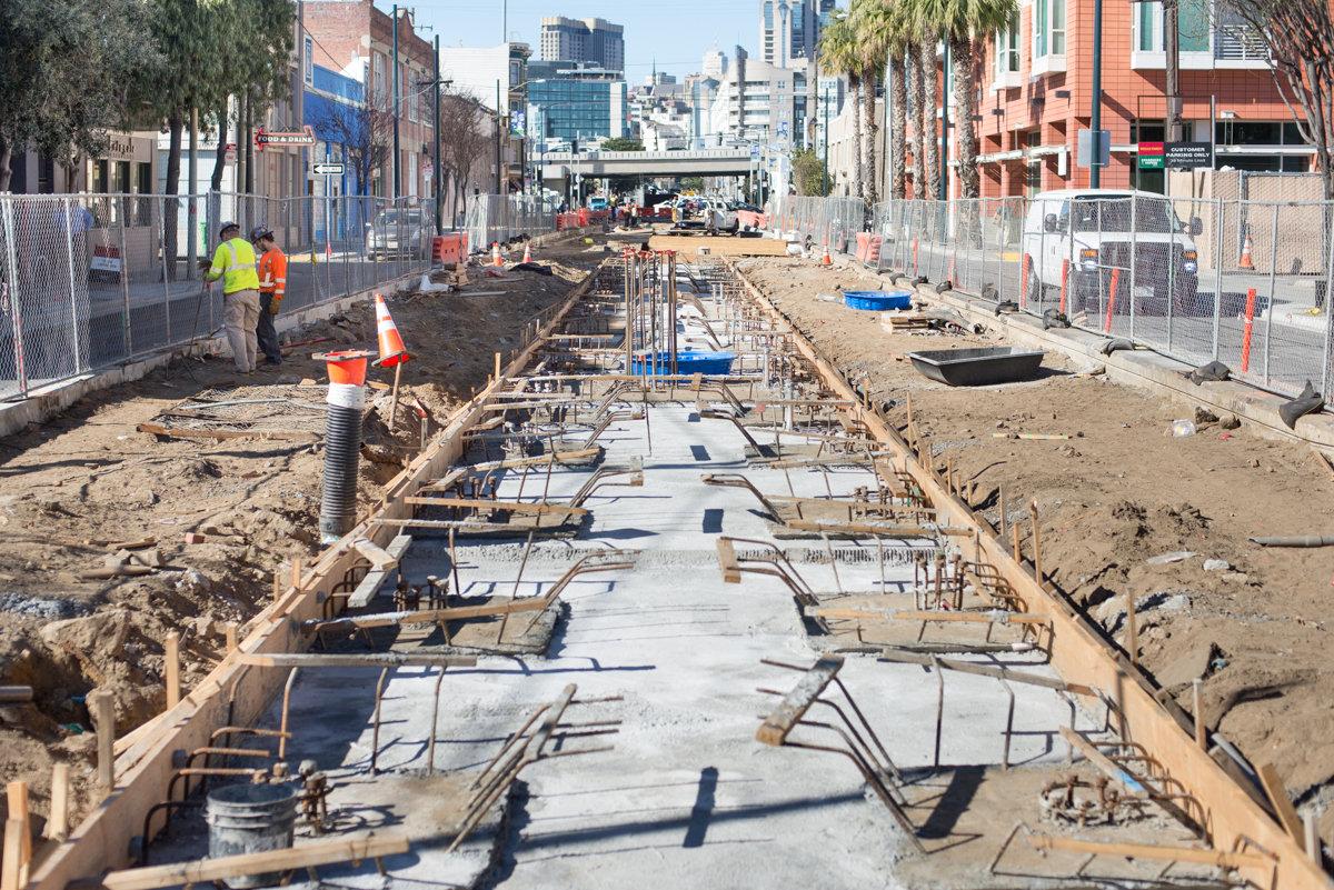 Construction site showing concrete foundation slab for subway station with steel reinforcement, workers, and city skyline in background.