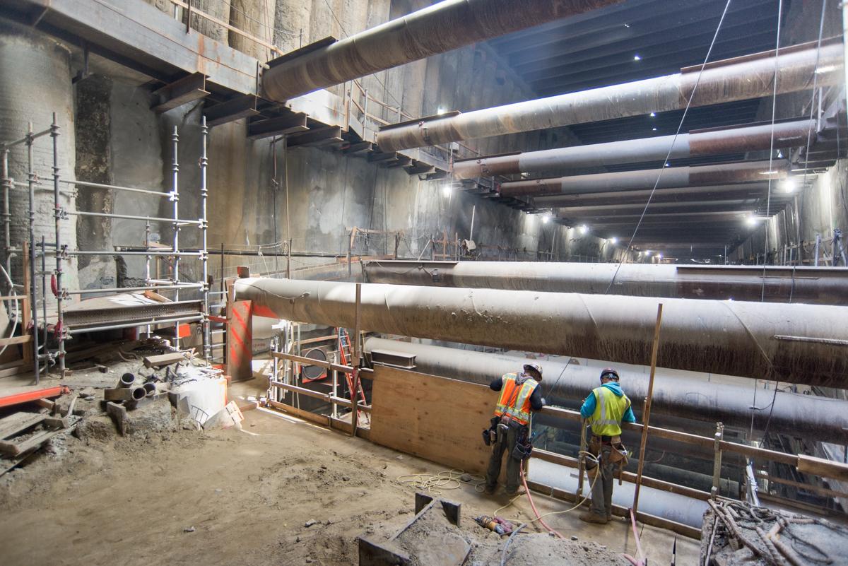 Workers in safety gear descend ladder into underground transit station construction site with large steel pipes and temporary bracing.
