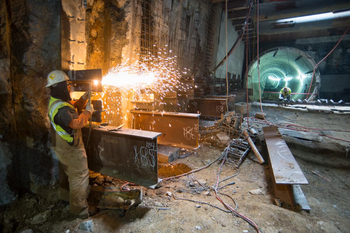 Worker in safety gear welding steel brackets, with sparks flying, inside an underground transit station tunnel.